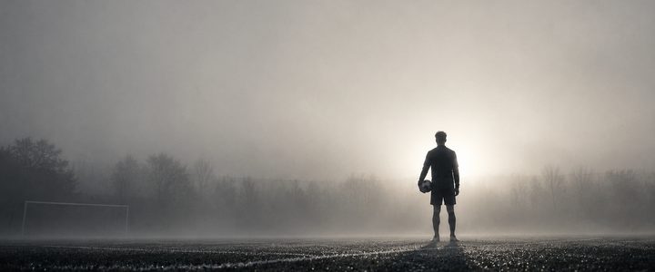 Un footballeur vu de dos, ballon sous le bras, seul sur un terrain dans le brouillard du matin.